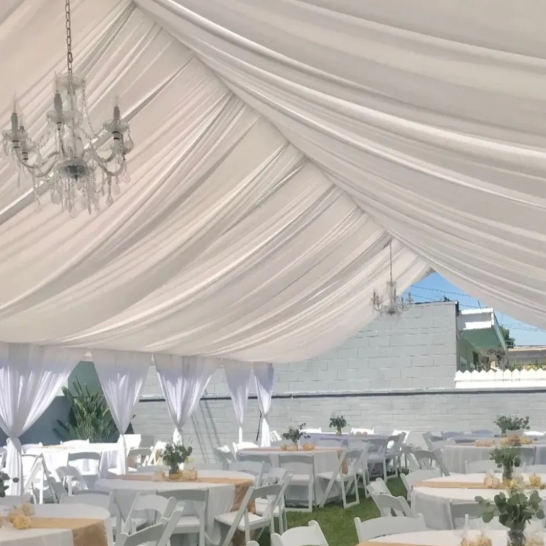 Elegant draped tent reception with crystal chandeliers, white folding chairs, and greenery centerpieces