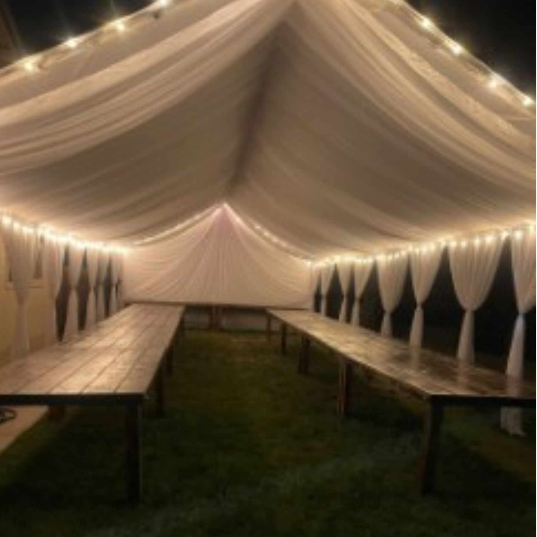 Illuminated draped tent with farm tables and string lights at night