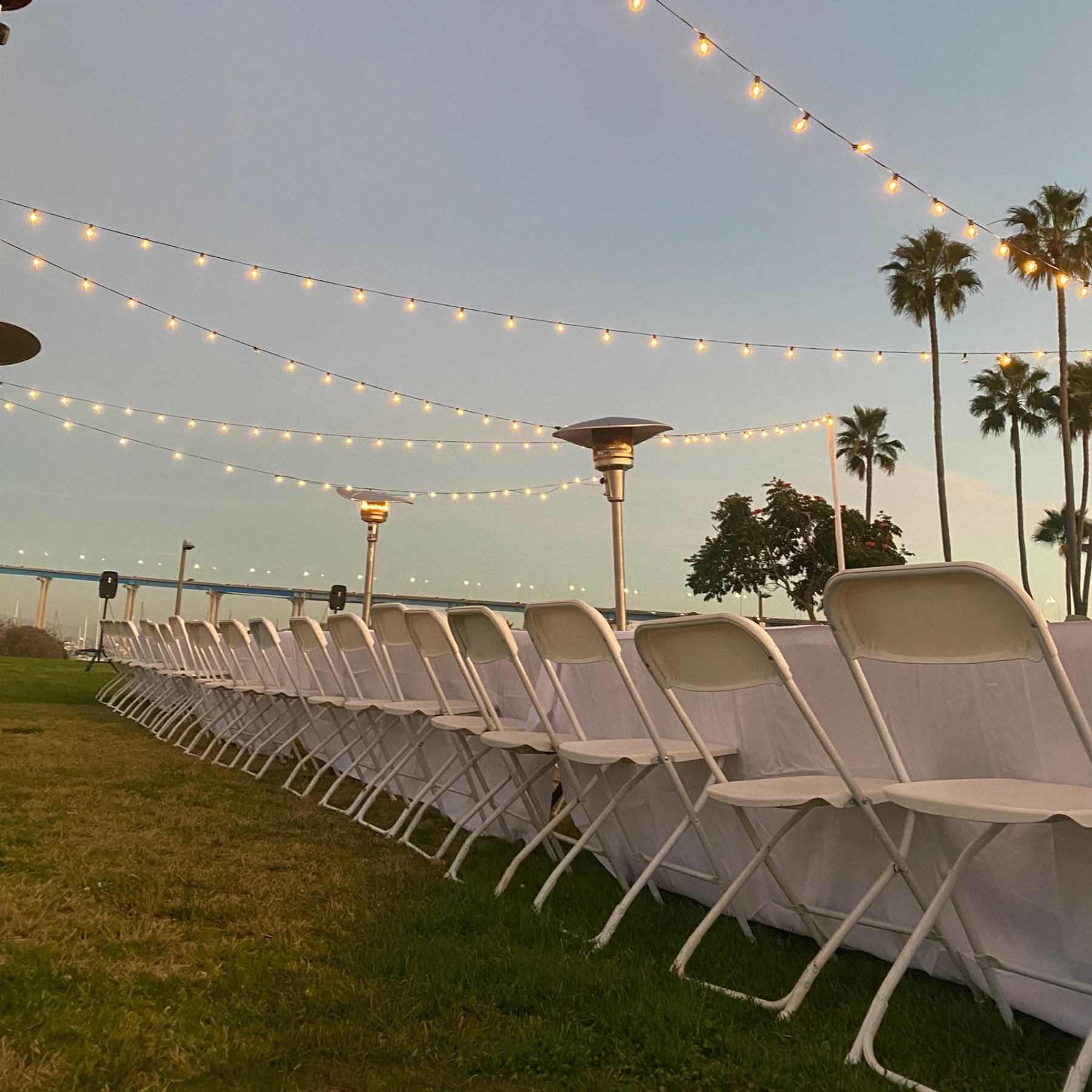 Outdoor event setup with string lights, patio heaters, and rows of white folding chairs