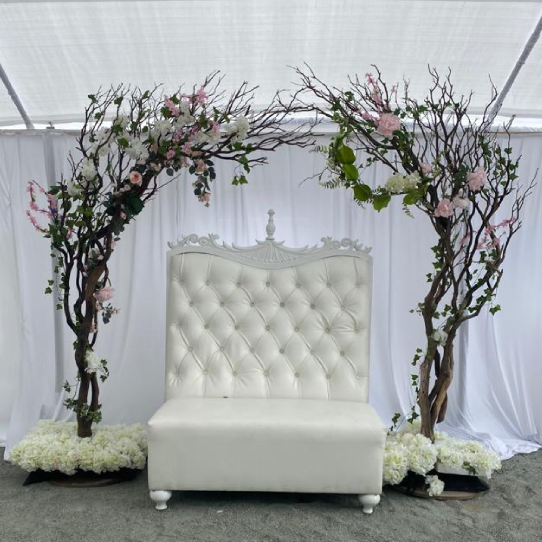 White tufted throne loveseat framed by a manzanita arch with pink and white florals under a draped tent